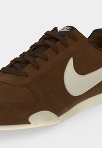 Καφέ Nubuck αθλητικό παπούτσι Nike με σκούρα καφέ κορδόνια και λευκό λογότυπο Nike swoosh στο πλάι, που δείχνει τις λεπτομέρειες μπροστά και στο πλάι.