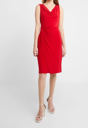 Una modelo viste un vestido rojo sin mangas con tejido drapeado, combinado con sandalias de tacón alto plateadas, todo lo cual se encuentra frente a un fondo blanco liso.