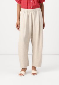 Beige linen trousers with a relaxed fit, featuring an elastic waistband and side pleats, paired with cream slide sandals.