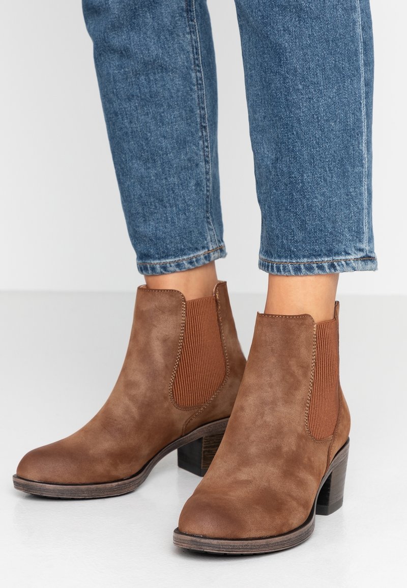 Brown suede ankle boots with elastic side panels and a stacked heel. The design features a rounded toe and textured accents.