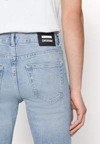 Svetlomodré džínsy majú vysoký pás, zadné vrecká na nášivky a čierny štítok s nápisom "DRDENIM" na páse.