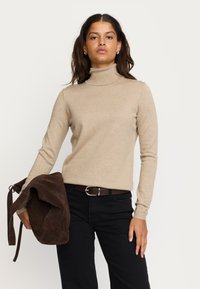 Beige Rollkragenpullover mit langen Ärmeln, ausgestattet mit einem gerippten Muster. Kombiniert mit schwarzen Hosen und einer braunen Wildleder-Schultertasche.