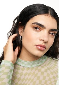 Top a righe verde e beige in tessuto trasparente con collo alto. Orecchini a cerchio argentati. Pelle liscia con trucco naturale, capelli scuri e mossi.