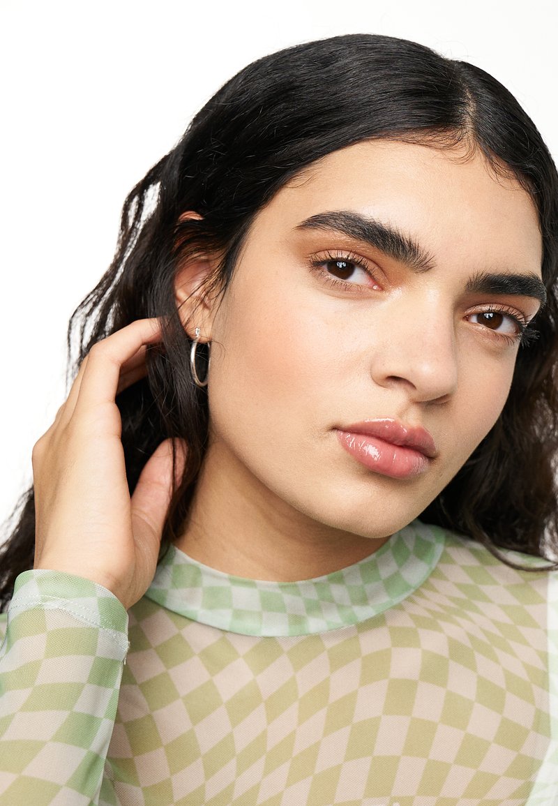 Top a righe verde e beige in tessuto trasparente con collo alto. Orecchini a cerchio argentati. Pelle liscia con trucco naturale, capelli scuri e mossi.