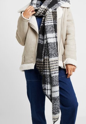 Personne portant une veste en peau lainée beige, une écharpe à carreaux noir et blanc, et un pantalon bleu foncé, ajustant son écharpe près du cou.