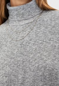 Pull à col roulé côtelé gris, doté d'un col montant et de colliers argentés superposés. Le tissu présente une finition texturée avec un motif subtil.