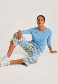 Lichtblauw langemouwen top gecombineerd met cropped broek met een ananaspatroon op een lichtblauwe achtergrond. Witte sneakers maken de look compleet.