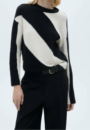 Femme portant un pull à rayures diagonales noir et blanc rentré dans un pantalon noir taille haute avec une ceinture noire.