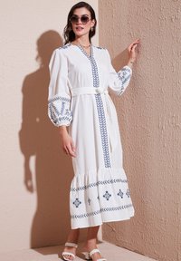 White maxi dress with balloon sleeves, navy embroidery, a v-neckline, and a tied waist belt; textured ruffled hem and sandals.