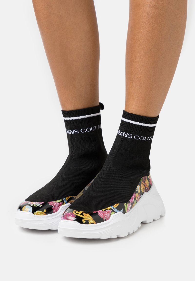 Jambes portant des baskets style chaussettes noires avec des semelles blanches et un motif baroque coloré sur les côtés et les orteils des chaussures, debout sur un fond blanc.