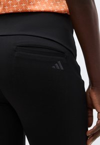 Μαύρο εφαρμοστό παντελόνι με πίσω τσέπη και διακριτικό λογότυπο Adidas, φορεμένο με πορτοκαλί εμπριμέ μπλούζα, που δείχνει το δεξί χέρι του φορέα στο πλάι.