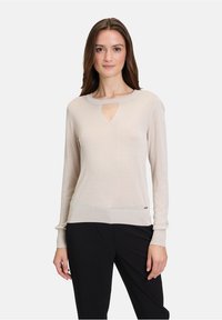 Pull en tricot beige clair avec un décolleté en forme de clé, des manches longues, des poignets côtelés et un ourlet ajusté. Agrémenté d'un petit logo.