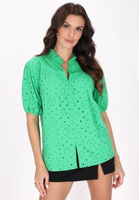 Grüne Bluse mit kurzen Ärmeln und Lochmuster, Knopfleiste und Faltenkragen. Weicher Stoff mit dezentem Glanz, kombiniert mit einem schwarzen Rock.