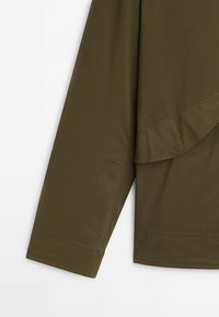 Manche et partie inférieure du dos d'une veste vert olive, montrant des coutures détaillées et un rabat en tissu à l'arrière.