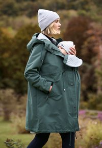 Vrouw met een groen jasje met baby in een draagzak, beiden met grijze gebreide mutsen, buiten in een natuurlijke omgeving met onscherpe bomen op de achtergrond.