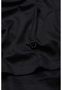 Schwarzes T-Shirt aus Stoff mit glatter Textur, ausgestattet mit einem kleinen, runden Etikett nahe dem Saum mit Details zur Marke. Dezente Nähte sichtbar.