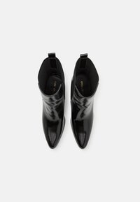 Paire de bottines noires brillantes à bout pointu avec languettes, vues de dessus sur un fond clair uni.