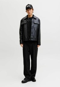 Joven de pie con chaqueta de cuero negra, suéter negro, pantalones de rayas finas, mocasines negros y una gorra negra con texto blanco.