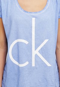Helles blaues Baumwoll-T-Shirt mit Rundhalsausschnitt und kurzen Ärmeln, mit einem großen weißen, gedruckten "ck"-Logo in der Mitte.