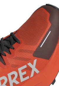 Oranžinės sportinės bateliai su tekstūruotu tinkleliu viršuje, juodais raišteliais ir ryškiu baltu logotipu. Turi paminkštintą pado dalį ir modernų dizainą.