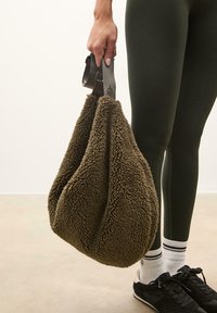 Bruine fuzzy tote bag gemaakt van textuurmateriaal met een brede opening aan de bovenkant, vastgehouden door een grijze verstelbare strap. Gebruikster draagt leggings en sneakers.