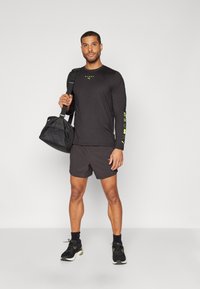 Schwarzes langärmliges Shirt mit gelben Akzenten und Logo, kombiniert mit dunklen Shorts. Das Model hält eine schwarze Sporttasche und trägt schwarze Sport Schuhe.