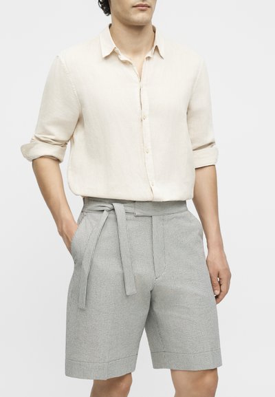 Άνδρας που φοράει μπεζ πουκάμισο με κουμπιά και γυρισμένα μανίκια και γκρι υφασμάτινες σορτς με υψηλή μέση, με υφασμάτινη ζώνη και τα χέρια στις τσέπες.