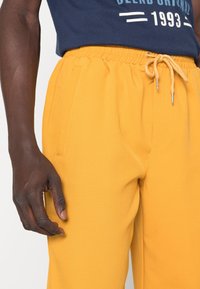 Osoba o ciemnej karnacji ubrana w żółte spodnie dresowe z sznurkiem i bocznymi kieszeniami, w połączeniu z granatową koszulką z napisem "1993" na wysokości talii.