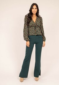 Blouse noire à fleurs avec des manches bouffantes, taille cintrée, associée à un pantalon évasé vert foncé et des bottes à talons beiges. Fond épuré.