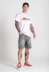 Bijela majica s crno-crvenim grafičkim uzorcima, sivi traper šortsevi s uzorkom i crvene tenisice s bijelim točkicama. Tetovaže vidljive na rukama i nogama.