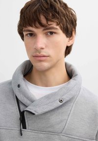 Sudadera de color gris claro con un cuello alto, cremallera y detalles de botones a presión, que presenta un tejido texturizado y un detalle de cremallera negra en contraste.