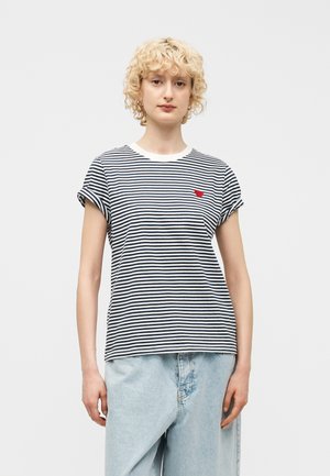 Blondynka o kręconych włosach, ubrana w czarno-białą paskowaną koszulkę z małym czerwonym sercem na piersi i jasnoniebieskie dżinsy.