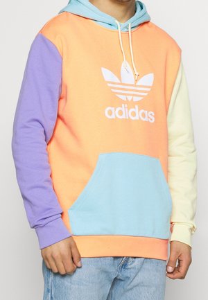 Muž nosí mikinu Adidas s barevnými bloky, levý rukáv fialový, pravý rukáv krémový, oranžový trup, světle modrá kapsa a šňůrky na kapuci.