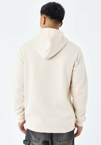 Sudadera beige de tela suave, con capucha ajustable, puños acanalados y un corte relajado. Las costuras visibles añaden textura y estructura.