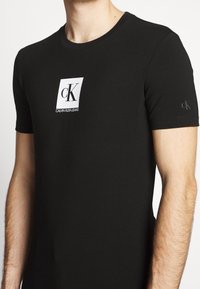 Camiseta negra de algodón con un logo rectangular blanco y texto en negro. Manga corta y diseño de cuello redondo. Logo sutil en la manga.