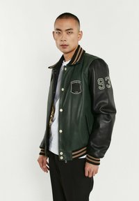 Veste universitaire en cuir avec un corps vert foncé et des manches noires, col et poignets en bord-côte rayés, patch avant et fermeture à bouton-pression.