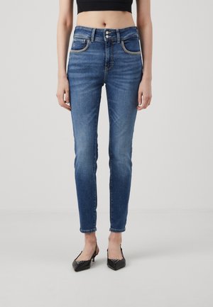 Kobieta nosząca niebieskie jeansy skinny z wysokim stanem i podwójnym zapięciem na guziki oraz czarne czółenka slingback z zaostrzonym noskiem, stoi na jednolitym tle.