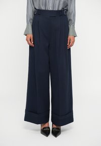 Pantalons navy à jambes larges avec une taille haute, présentant des plis à l'avant et des détails de boucle. Le tissu a l'air lisse avec une conception structurée.