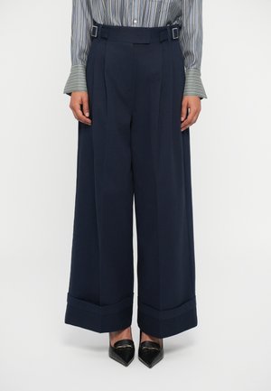 Pantaloni navy a gamba ampia con vita alta, caratterizzati da pieghe frontali e dettagli con fibbia. Il tessuto appare liscio con un design strutturato.