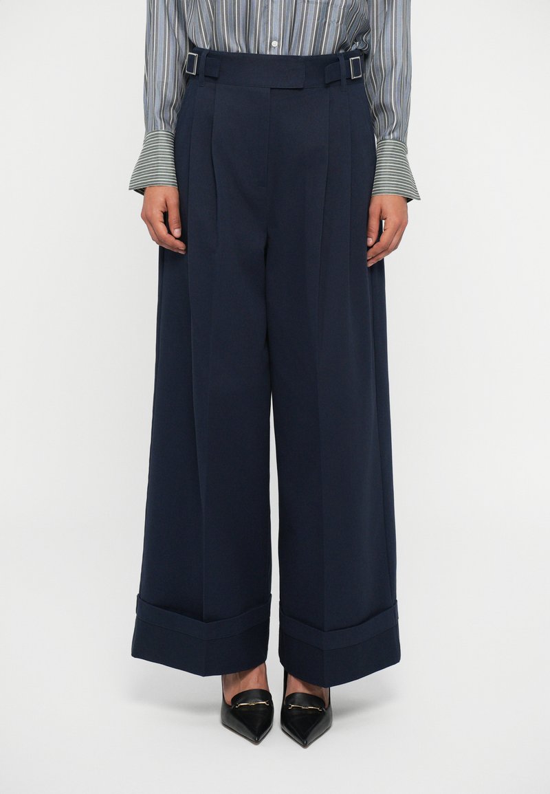 Pantalons navy à jambes larges avec une taille haute, présentant des plis à l'avant et des détails de boucle. Le tissu a l'air lisse avec une conception structurée.