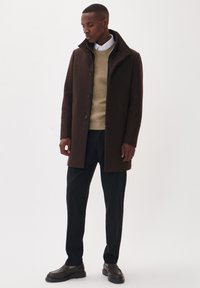 Man draagt een bruine jas met knopen over een beige trui, wit overhemd, zwarte broek en donkerbruine loafers, staand tegen een witte achtergrond.