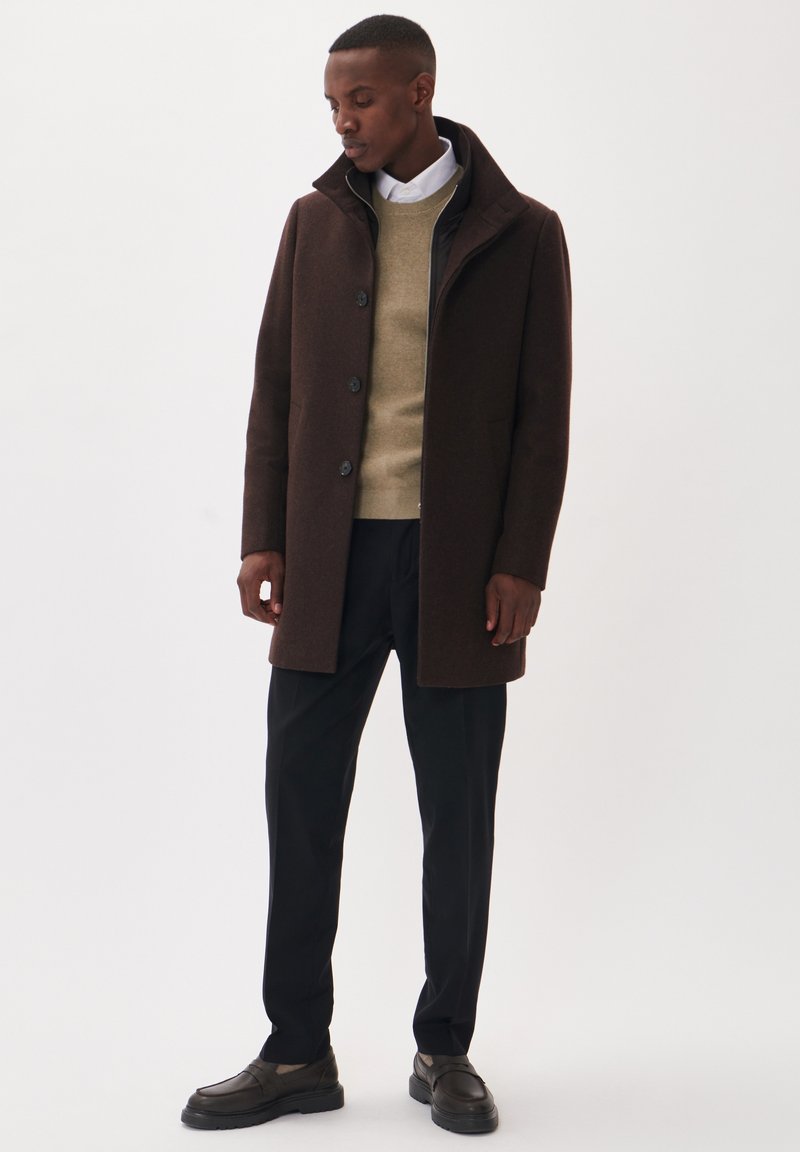 Man draagt een bruine jas met knopen over een beige trui, wit overhemd, zwarte broek en donkerbruine loafers, staand tegen een witte achtergrond.