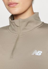Haut de sport beige avec un col montant, fermeture éclair demi-longueur, et logo blanc. Texture lisse sans motifs ni embellissements visibles.