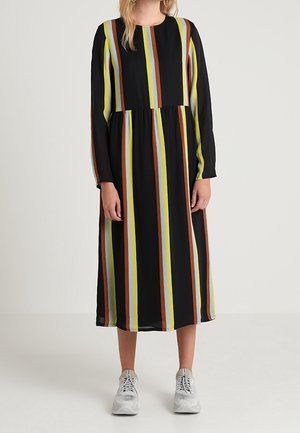 Mujer que lleva un vestido midi negro de manga larga con rayas verticales en amarillo, marrón y azul claro, combinado con zapatillas blancas.