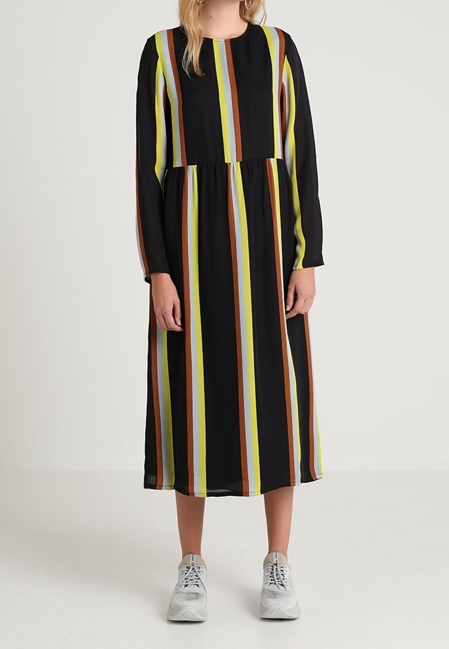 Femme portant une robe midi noire à manches longues avec des rayures verticales jaunes, marron et bleu clair, associée à des baskets blanches.