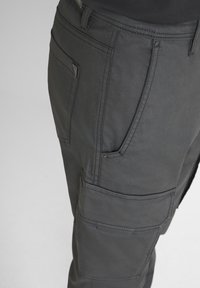 Primo piano di pantaloni cargo grigio scuro che mostrano le tasche frontali e laterali con dettagli di cucitura su uno sfondo neutro.