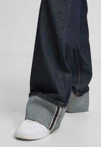 Plati kelnės iš tamsiai mėlynos džinsinės medžiagos su kontrastiniu pilku atvartėliu. Yra baltos siūlės ir plyšio detalė, derinamos su baltais sportbačiais.