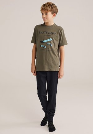 Garçon debout portant un t-shirt graphique skateboard vert olive, pantalon noir et chaussettes noires sur fond uni.