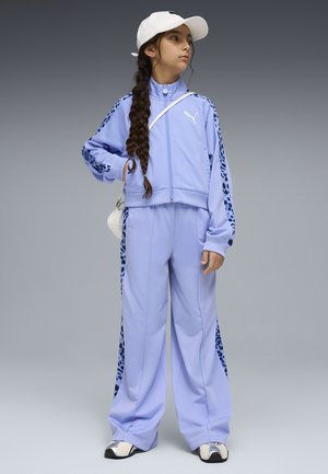 Jong meisje met lange vlecht, gekleed in een lila Puma trainingspak met luipaardprintstrepen, witte pet, schoudertas en sneakers, staand en naar rechts kijkend.