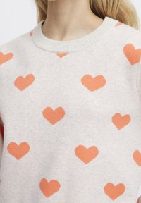 Pull gris clair avec un motif intégral de cœurs orange. Col rond côtelé et texture douce en maille.